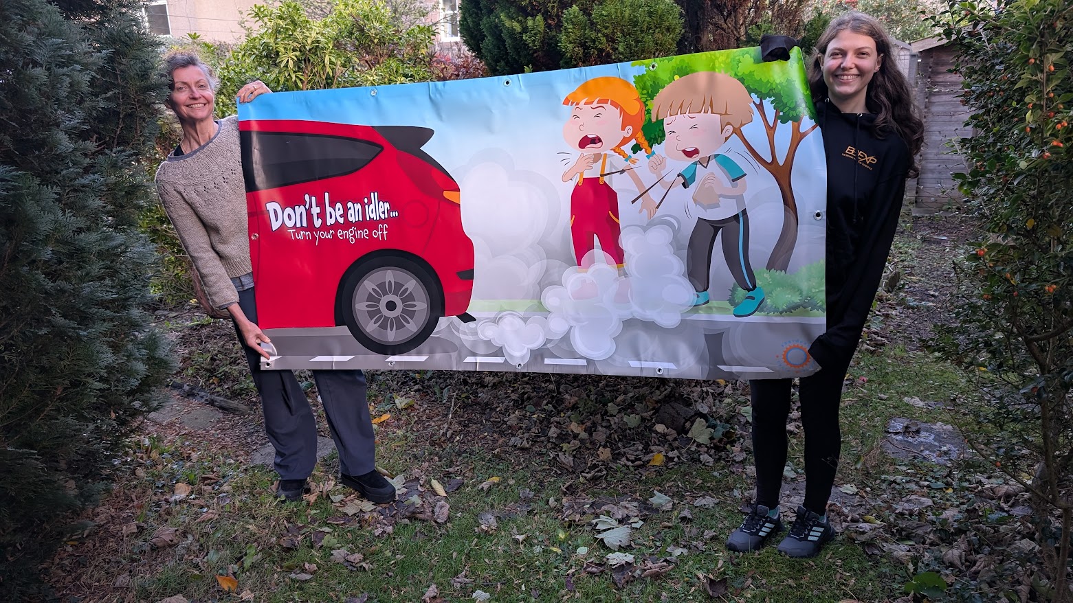 Don't be an idler banner showing two children coughing while inhaling fumes coming from a red car