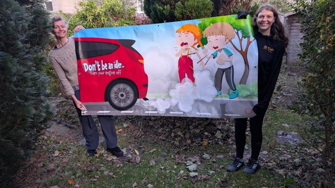 Don't be an idler banner showing two children coughing while inhaling fumes coming from a red car